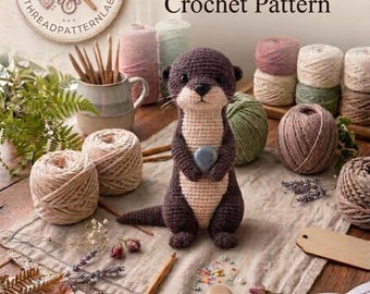 May include: A crocheted dark brown and beige river otter holding a small gray stone, surrounded by various yarn balls, wooden crochet hooks, dried flowers, and a pattern book with the text The Little River Otter Crochet Pattern and INSTANT PDF DOWNLOAD.
