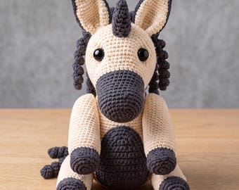 Könnte beinhalten: Ein gehäkeltes Einhorn mit cremefarbenem Körper, dunkelgrauen Hufen, Schnauze, Horn und lockiger Mähne. Der Text Cozy Curls Crochet Unicorn Pattern und Instant PDF Download ist sichtbar. Das Einhorn hat große schwarze Augen und sitzt.