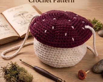 May include: A crochet mini bag shaped like a mushroom with a dark maroon cap featuring white stitched spots and a cream-colored stem. The bag has a cream-colored cord strap. Text reads: Magic Mushroom Mini Bag Crochet Pattern. Instant PDF Download.