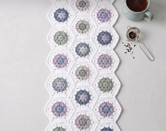 Könnte beinhalten: Ein weißer Häkel-Tischläufer aus miteinander verbundenen sechseckigen Motiven, die jeweils ein zentrales Blumenmuster in Salbeigrün, Staubblau und Lavendel aufweisen. Der Text "Boho Floral Hexagon Table Runner Crochet Pattern" und "Instant PDF Download" ist sichtbar.