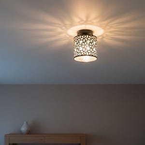 May include: A ceiling-mounted light fixture with a cylindrical shade. The shade is white with a floral pattern, and the light casts a starburst pattern on the ceiling. The fixture has a metallic finish.