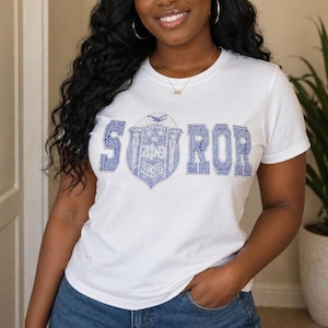 May include: White t-shirt with the word "SOROR" in blue rhinestones. The letter "S" is followed by a shield design with stars and a banner. The shirt is short-sleeved and made of a soft material.