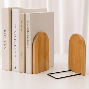 Puede incluir: Dos sujetalibros de bambú con acabado de vetas de madera natural. Los sujetalibros tienen una parte superior curvada y una base de metal negro. Se ven libros con los títulos "KINFOLK", "THE KINFOLK HOME" y "Beyond the Landscape".