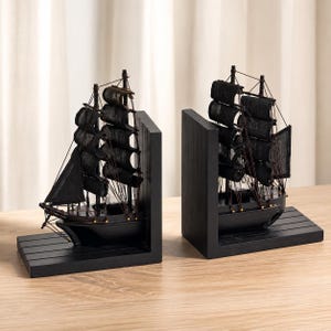 Puede incluir: Un par de sujetalibros de madera negra, cada uno con un modelo detallado de un velero con velas de tela negra, finas líneas de aparejo y una base resistente en forma de L. Los barcos están colocados de manera que miran hacia el interior.