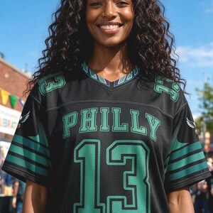 May include: A black V-neck jersey featuring the word PHILLY in green letters above the number 13 in large green font. The sleeves have green stripes and small eagle graphics, with the number 13 repeated on the shoulders in green.