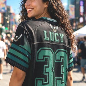 May include: A black sports jersey featuring the name LUCY in green letters above the number 33 in large green font. The sleeves have green and black stripes with an eagle emblem on the shoulder. The collar displays the text Philadelphia.