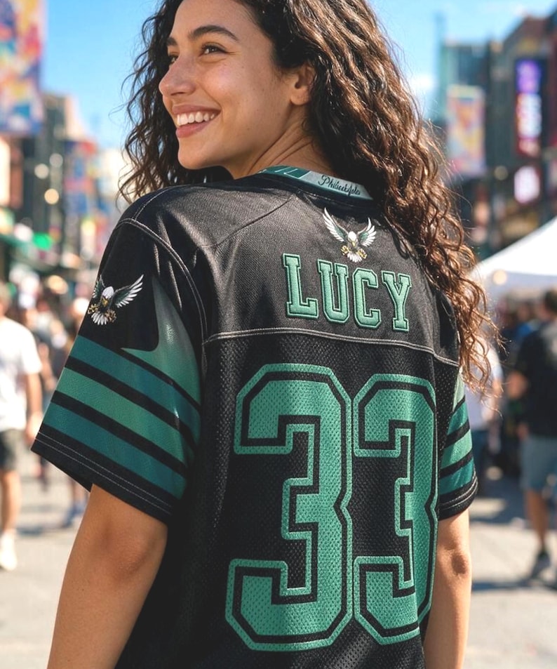 May include: A black sports jersey featuring the name LUCY in green letters above the number 33 in large green font. The sleeves have green and black stripes with an eagle emblem on the shoulder. The collar displays the text Philadelphia.