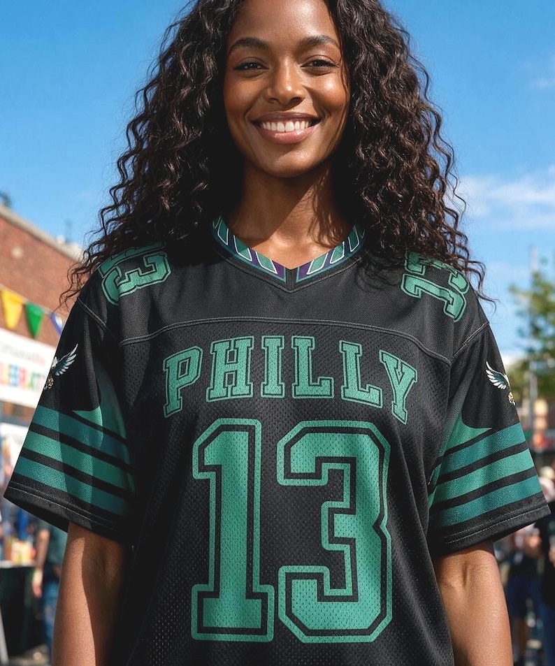 May include: A black V-neck jersey featuring the word PHILLY in green letters above the number 13 in large green font. The sleeves have green stripes and small eagle graphics, with the number 13 repeated on the shoulders in green.