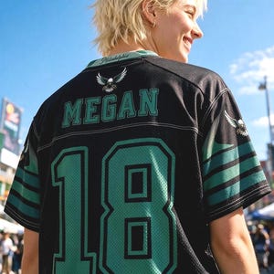 May include: A black sports jersey featuring the name MEGAN and the number 18 in large teal lettering with black outlines. The shirt includes a small eagle graphic, the word Philadelphia on the collar, and teal and black striped sleeve accents.