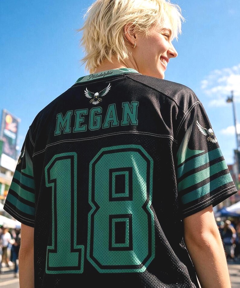 May include: A black sports jersey featuring the name MEGAN and the number 18 in large teal lettering with black outlines. The shirt includes a small eagle graphic, the word Philadelphia on the collar, and teal and black striped sleeve accents.