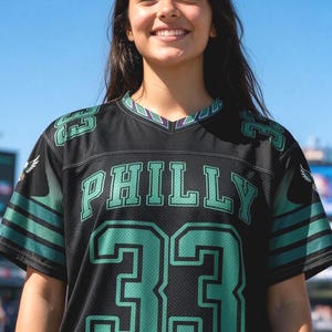 May include: A black athletic jersey with a V-neck collar featuring the word PHILLY in green letters across the chest and the number 33 in large green digits below. The sleeves have green stripes and the number 33 is printed on each shoulder.