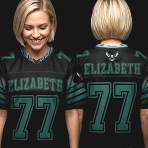 May include: A black jersey featuring the name ELIZABETH and the number 77 in green lettering on the front and back. The jersey includes a V-neck collar with green and white accents, green sleeve stripes, and an eagle emblem on the back neckline.