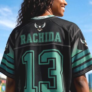 May include: A black sports jersey featuring the name RACHIDA and the number 13 in green with black outlines. The back of the jersey displays a small eagle graphic below the collar, which is printed with the word Philadelphia in white script.