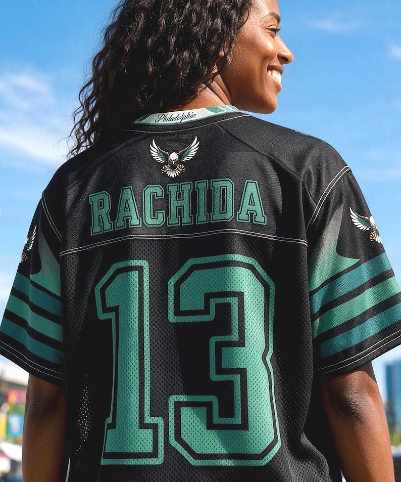 May include: A black sports jersey featuring the name RACHIDA and the number 13 in green with black outlines. The back of the jersey displays a small eagle graphic below the collar, which is printed with the word Philadelphia in white script.