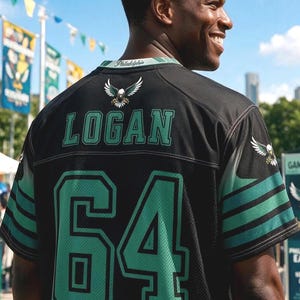 May include: A black sports jersey featuring the name LOGAN and the number 64 in green with black outlines on the back. The jersey includes an eagle graphic on the upper back and sleeves, with the word Philadelphia printed on the collar trim.