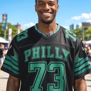May include: A black short-sleeved jersey with a V-neck collar featuring green trim. The front displays the word PHILLY in green letters above the large number 73 in green. The number 73 is also on each shoulder, and green stripes accent the sleeves.