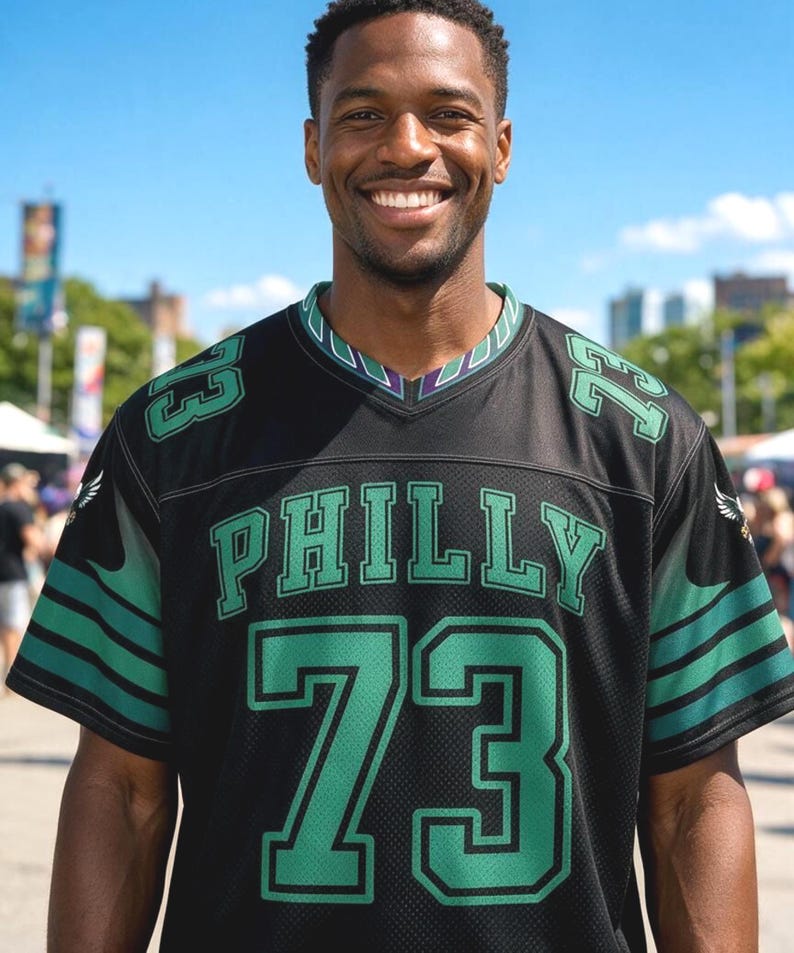 May include: A black short-sleeved jersey with a V-neck collar featuring green trim. The front displays the word PHILLY in green letters above the large number 73 in green. The number 73 is also on each shoulder, and green stripes accent the sleeves.