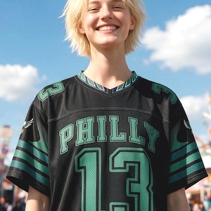 May include: A black short-sleeved sports jersey featuring the word PHILLY in green letters above the number 13 in large green font. The sleeves have green and black stripes with small eagle graphics, and the V-neck collar has green trim.