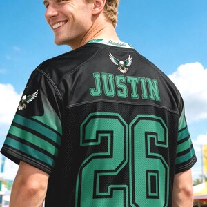 May include: A black sports jersey featuring the name JUSTIN and the number 26 in green with black outlines. The back of the jersey displays an eagle emblem, green and black sleeve stripes, and the word Philadelphia printed on the inner collar.