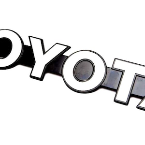 Puede incluir: Un emblema de Toyota en blanco y negro con el nombre de la marca en letras blancas en negrita. El emblema está ligeramente inclinado sobre un fondo blanco. Las letras son gruesas y estilizadas.