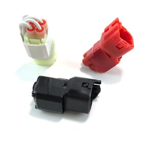 Puede incluir: Se muestran tres conectores eléctricos. Uno es rojo, otro negro y el tercero blanco y amarillo. Los conectores son de plástico y tienen varias formas y tamaños. Es probable que estos conectores se utilicen en aplicaciones automotrices o electrónicas.