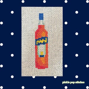 May include: A needlepoint canvas featuring a stitched design of an orange bottle with a blue cap and a blue label containing yellow text. The bottle has a light blue outline and a small yellow detail near the base, all on a white grid mesh.