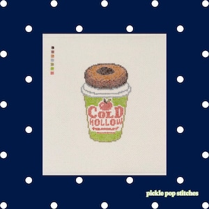 May include: A cross-stitch pattern on white fabric featuring a green cup with a brown donut on top. The cup displays a red apple graphic and the text COLD HOLLOW in red letters. A small vertical color key is visible on the upper left side of the design.