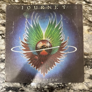May include: A square vinyl record cover for the album Evolution by the band Journey. The cover features a central dark sphere with a landscape reflection, surrounded by large, stylized green and brown wings and a thin white ring against a blue background.