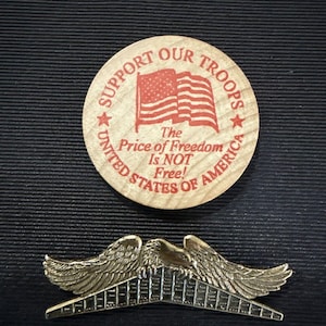 May include: A wooden round with the text "SUPPORT OUR TROOPS" and an American flag graphic. Below, it reads "The Price of Freedom Is NOT Free! UNITED STATES OF AMERICA." Below is a gold-colored metal pin with eagle wings.