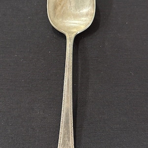Puede incluir: Una cuchara de metal plateado con un cuenco ovalado alargado y un mango delgado. El mango está decorado con un delicado patrón geométrico repetitivo en los bordes y un pequeño motivo grabado intrincado en la base del tallo.