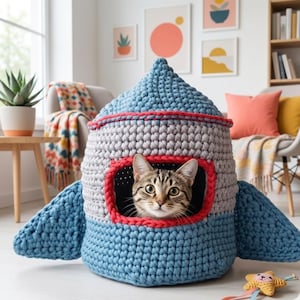 May include: A handmade, crochet cat bed shaped like a rocket ship. The bed is primarily blue, gray, and red, with a circular window framing a tabby cat. The rocket has wing-like structures on the sides.