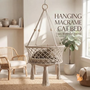 May include: A hanging macrame cat bed featuring a circular woven base with a soft cushion, suspended by knotted cotton cords and wooden beads. The text reads HANGING MACRAME CAT BED Cozy Boho Cat Swing. A cat is resting inside the beige woven structure.