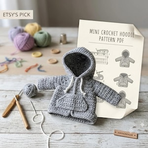 May include: A gray crocheted mini hoodie with a front pocket and white drawstring, accompanied by a PDF pattern sheet titled MINI CROCHET HOODIE PATTERN PDF, two wooden crochet hooks, a small ball of gray yarn, and a small wooden tag labeled HANDMADE.