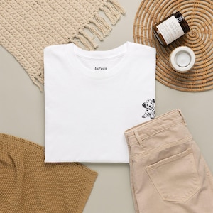 Può includere: Una t-shirt bianca a maniche corte con una piccola illustrazione in bianco e nero di un cucciolo di dalmata sul lato inferiore destro. Il colletto interno mostra il testo laFras in lettere nere. La maglietta è abbinata a pantaloni in denim beige.
