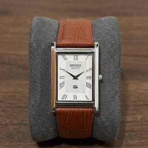 Puede incluir: Un reloj Seiko Quartz rectangular con caja plateada y correa de cuero marrón. La esfera del reloj es blanca con números romanos y agujas negras. El reloj se muestra sobre un cojín gris.