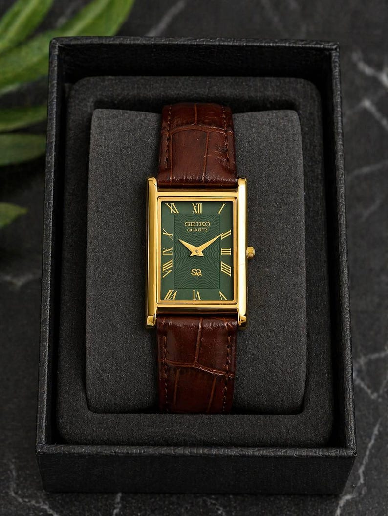 Puede incluir: Un reloj rectangular Seiko Quartz con caja dorada y esfera verde oscuro con n&uacute;meros romanos dorados. El reloj cuenta con una correa de cuero marr&oacute;n con textura de cocodrilo y se presenta en una caja de exhibici&oacute;n de color negro.