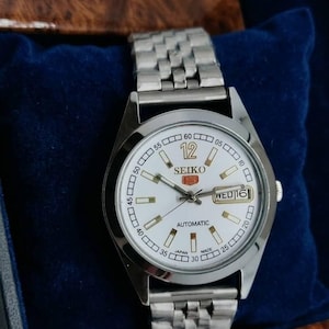 Puede incluir: Un reloj automático Seiko plateado con esfera blanca, números y marcadores negros. El reloj muestra el día y la fecha, con una correa plateada. El reloj se presenta en una caja forrada de terciopelo azul.