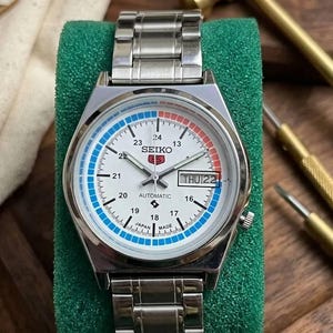Op de afbeelding: Een zilverkleurig Seiko automatisch polshorloge met een witte wijzerplaat, met blauwe en rode accenten. Het horloge geeft de dag en datum weer, met de tekst "SEIKO" en "AUTOMATIC" zichtbaar. Het horloge ligt op een groene ondergrond.