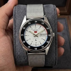 Op de afbeelding: Een zilveren Seiko 5 GMT automatische horloge met een witte wijzerplaat, zwarte en rode accenten en een mesh band. Het horloge toont de dag en datum. Het horloge wordt gepresenteerd op een grijs fluwelen kussen.