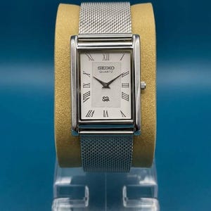 Peut inclure: Une montre Seiko Quartz rectangulaire avec un bracelet en maille argentée. Le cadran de la montre est blanc avec des chiffres romains et des aiguilles noirs. La montre est présentée sur un support beige sur fond bleu.