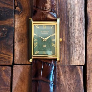 Puede incluir: Un reloj de pulsera rectangular Seiko Quartz con caja dorada, esfera verde con patr&oacute;n de ondas conc&eacute;ntricas y n&uacute;meros romanos dorados. El reloj cuenta con una correa de cuero marr&oacute;n oscuro con un acabado texturizado de estilo cocodrilo.