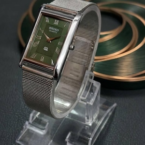 Puede incluir: Un reloj Seiko Quartz rectangular con esfera verde y correa de malla plateada. La esfera del reloj presenta números romanos y una manecilla dorada. El reloj se exhibe sobre un soporte de plástico transparente.