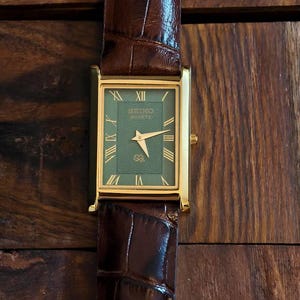 Puede incluir: Un reloj rectangular Seiko Quartz con caja en tono dorado y esfera verde oscuro con un patr&oacute;n de ondas texturizado. El reloj cuenta con marcadores de hora en n&uacute;meros romanos dorados, manecillas doradas y una correa de cuero marr&oacute;n.