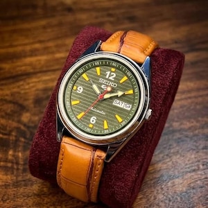 Pode incluir: Um relógio de pulso automático Seiko com mostrador verde, caixa prateada e bracelete de couro castanho. O mostrador mostra o dia e a data. O relógio está sobre uma superfície de veludo bordô.