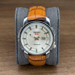 Puede incluir: Reloj de pulsera automático Seiko con esfera plateada, visualización de día y fecha, y correa de cuero marrón. El reloj tiene una caja de acero inoxidable y se muestra sobre un cojín gris.