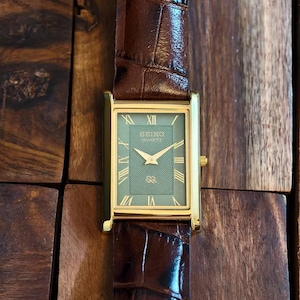 Puede incluir: Un reloj de pulsera rectangular Seiko Quartz con caja dorada, esfera verde con patr&oacute;n de ondas conc&eacute;ntricas, manecillas doradas e &iacute;ndices de n&uacute;meros romanos. El reloj cuenta con una correa de cuero marr&oacute;n oscuro con textura de cocodrilo.