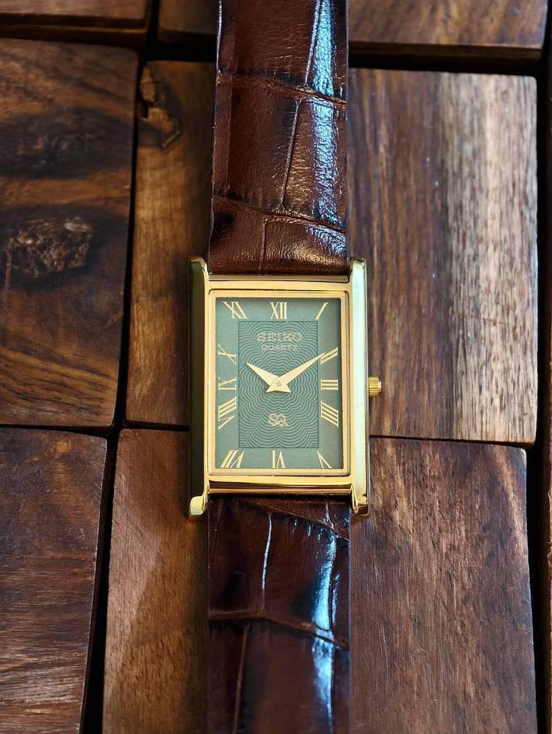 Puede incluir: Un reloj de pulsera rectangular Seiko Quartz con caja dorada, esfera verde con patr&oacute;n de ondas conc&eacute;ntricas, manecillas doradas e &iacute;ndices de n&uacute;meros romanos. El reloj cuenta con una correa de cuero marr&oacute;n oscuro con textura de cocodrilo.