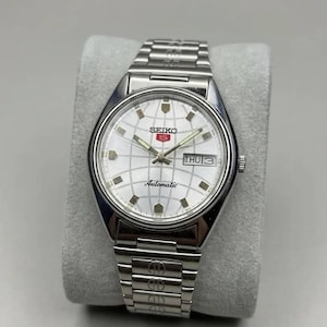 May include: Silver Seiko 5 Automatic wristwatch with a white grid-patterned dial. The watch has a day-date display and a stainless steel strap. The watch is displayed on a grey cushion.