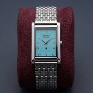 May include: A rectangular Seiko Quartz watch with a silver band and a turquoise face. The watch has Roman numerals and silver hands. The watch is displayed on a burgundy velvet surface.