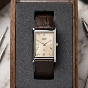 Könnte beinhalten: Eine rechteckige Seiko Quartz-Uhr mit silbernem Gehäuse und braunem Lederarmband, präsentiert in einer Holzbox. Das Zifferblatt der Uhr ist cremefarben mit römischen Ziffern und dem Seiko-Logo. Das Armband hat ein Krokodilmuster.
